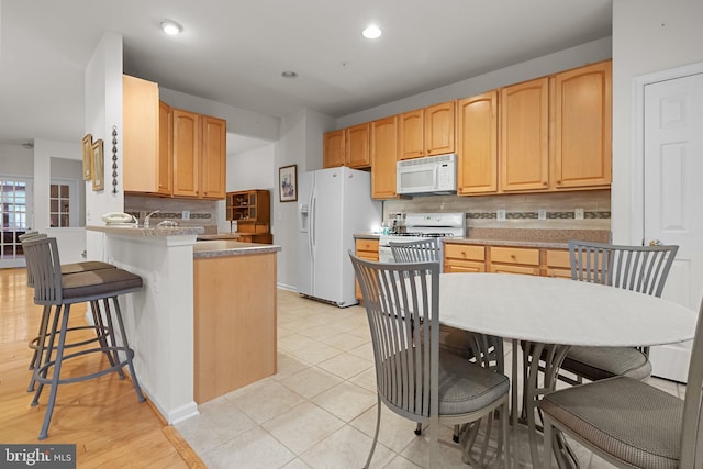 kitchen with white appliances, decorative backsplash, light brown cabinetry, a kitchen bar, and kitchen peninsula