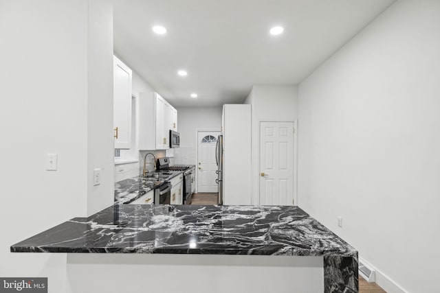 kitchen featuring kitchen peninsula, backsplash, stainless steel appliances, dark stone countertops, and white cabinetry