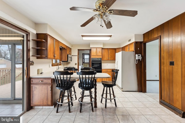 kitchen featuring a breakfast bar, black double oven, wooden walls, ceiling fan, and white fridge