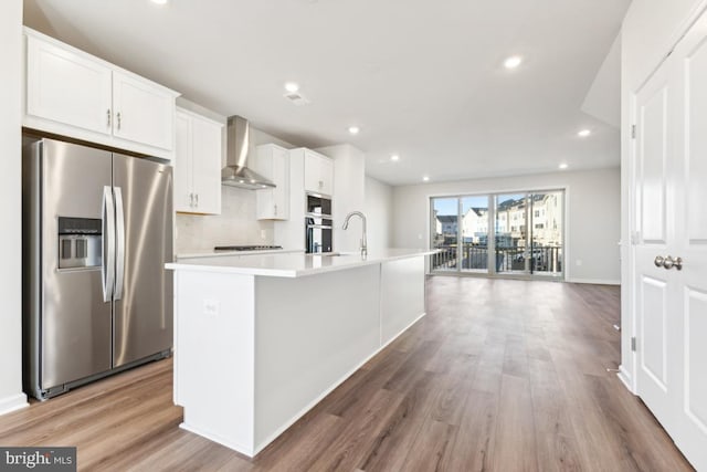 kitchen featuring appliances with stainless steel finishes, wall chimney exhaust hood, hardwood / wood-style flooring, white cabinetry, and an island with sink