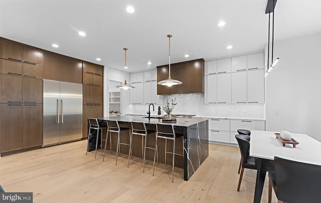 kitchen with stainless steel built in fridge, an island with sink, hanging light fixtures, and light hardwood / wood-style flooring