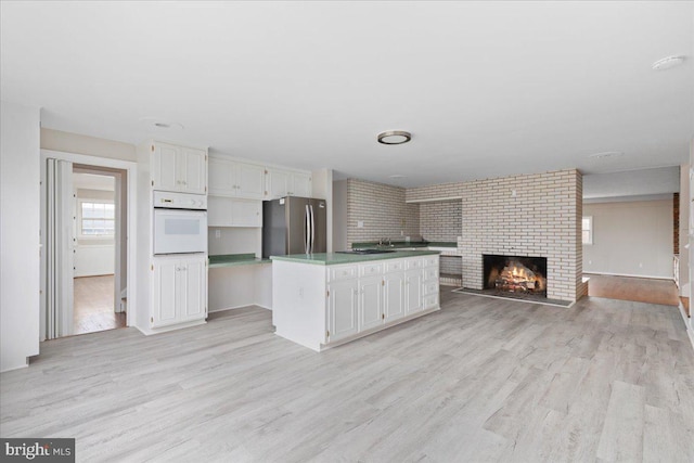 kitchen with white oven, white cabinets, a brick fireplace, light wood-type flooring, and stainless steel refrigerator