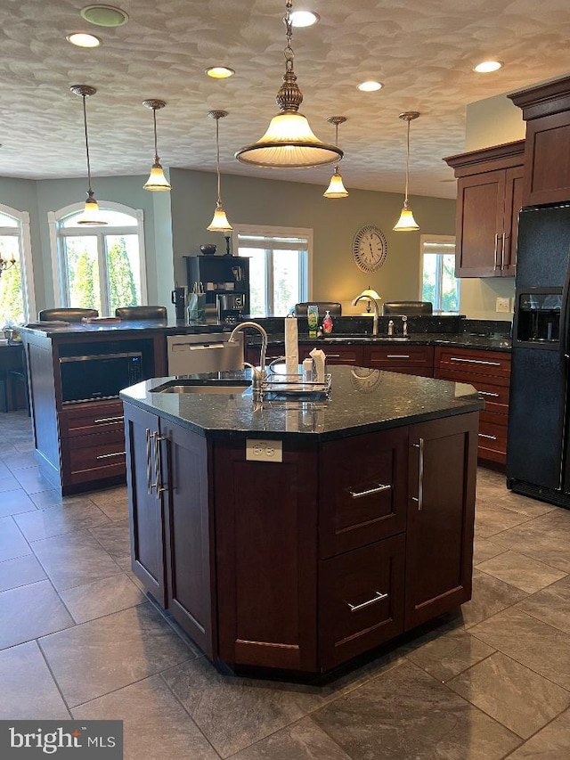 kitchen with dark stone counters, black fridge with ice dispenser, hanging light fixtures, and an island with sink