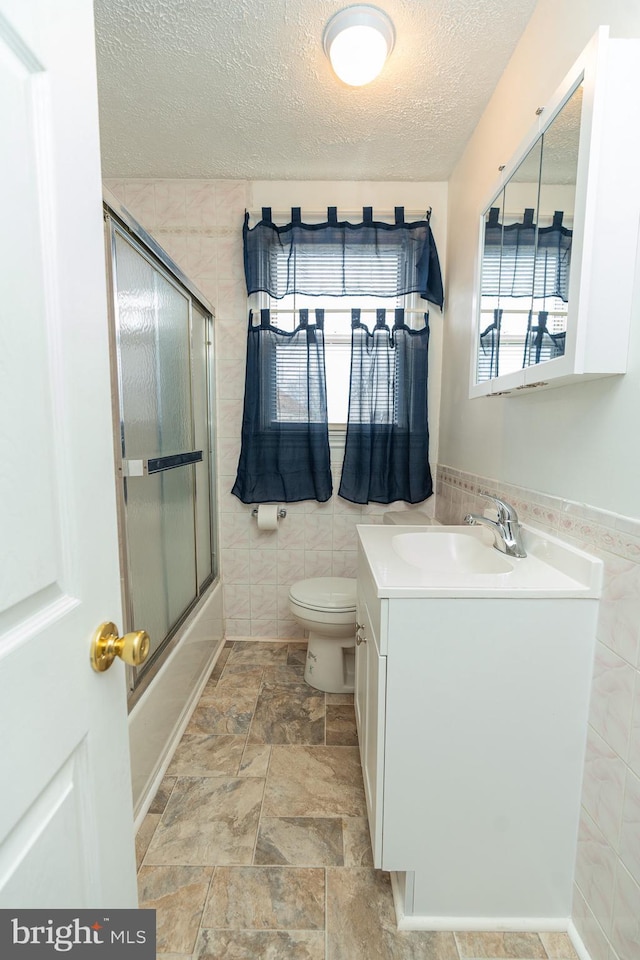 full bathroom with vanity, shower / bath combination with glass door, toilet, a textured ceiling, and tile walls