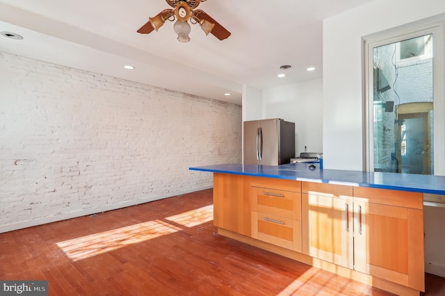 kitchen featuring stainless steel refrigerator, ceiling fan, brick wall, hardwood / wood-style floors, and light brown cabinetry