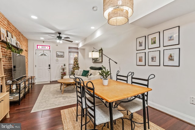 dining space with brick wall, dark wood-type flooring, and ceiling fan with notable chandelier