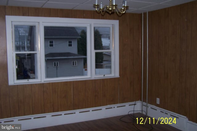 empty room featuring a chandelier, a baseboard radiator, a drop ceiling, and wood walls