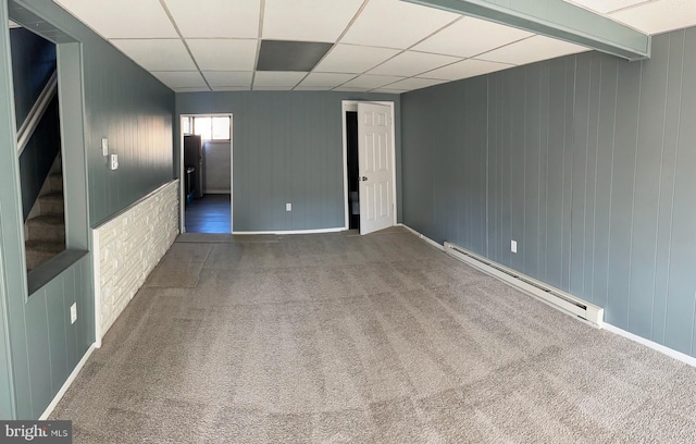 carpeted empty room featuring wooden walls, a drop ceiling, and a baseboard heating unit