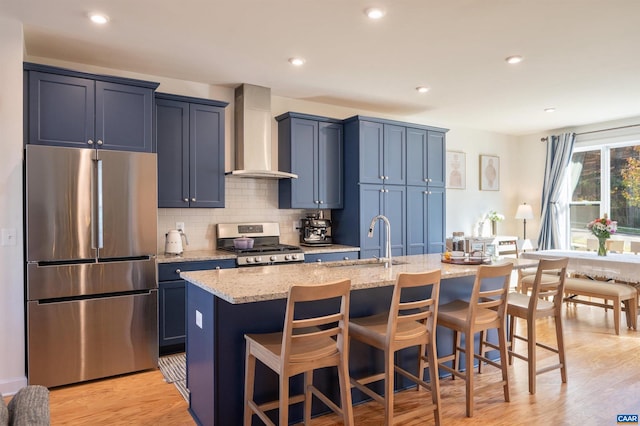 kitchen featuring wall chimney exhaust hood, stainless steel appliances, an island with sink, light hardwood / wood-style floors, and a kitchen bar