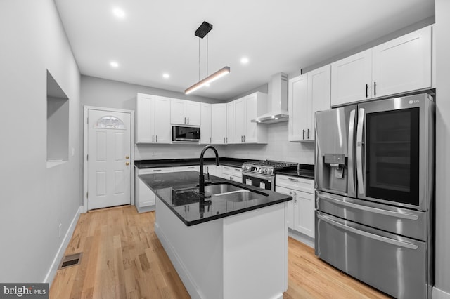 kitchen featuring white cabinets, wall chimney range hood, light hardwood / wood-style flooring, an island with sink, and stainless steel appliances