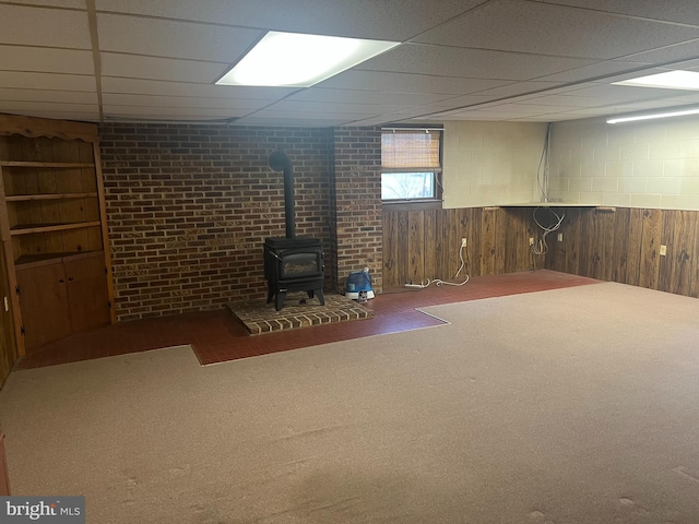 basement with carpet flooring, a wood stove, wood walls, and a drop ceiling