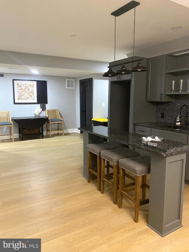 kitchen with light wood-type flooring, backsplash, dark stone countertops, hanging light fixtures, and a breakfast bar area