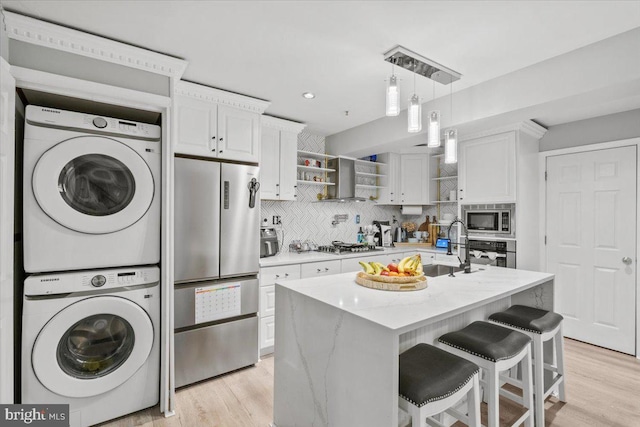 kitchen with stacked washing maching and dryer, stainless steel appliances, a center island with sink, light hardwood / wood-style floors, and white cabinetry