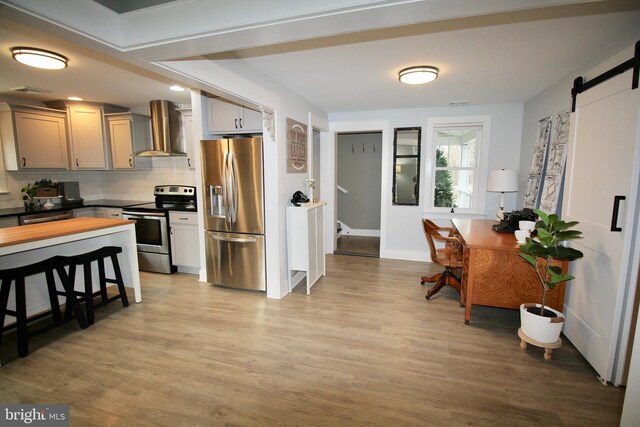 kitchen featuring gray cabinetry, stainless steel appliances, wall chimney range hood, a barn door, and light hardwood / wood-style flooring