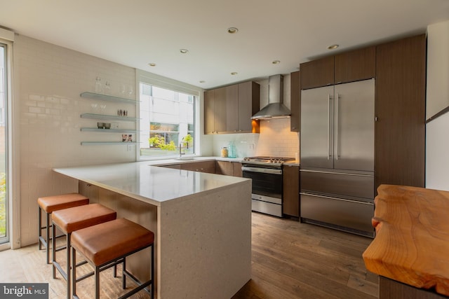 kitchen with a kitchen breakfast bar, appliances with stainless steel finishes, light wood-type flooring, kitchen peninsula, and wall chimney range hood