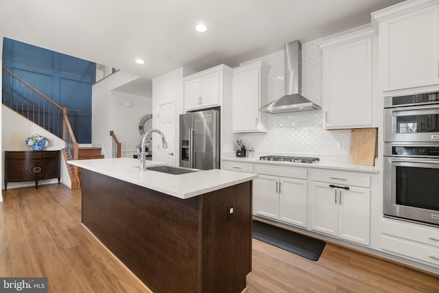 kitchen with sink, wall chimney range hood, a kitchen island with sink, white cabinets, and appliances with stainless steel finishes