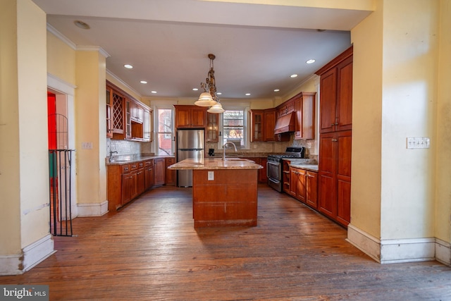 kitchen with dark wood-type flooring, custom exhaust hood, hanging light fixtures, a center island with sink, and appliances with stainless steel finishes