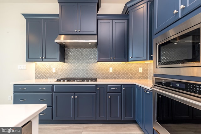 kitchen featuring under cabinet range hood, blue cabinetry, backsplash, appliances with stainless steel finishes, and light countertops