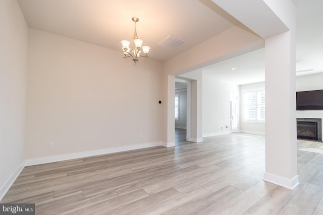 spare room with light wood finished floors, visible vents, baseboards, a glass covered fireplace, and a notable chandelier