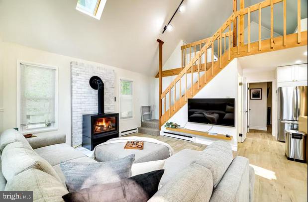 living room with a wood stove, a skylight, baseboard heating, high vaulted ceiling, and light hardwood / wood-style floors