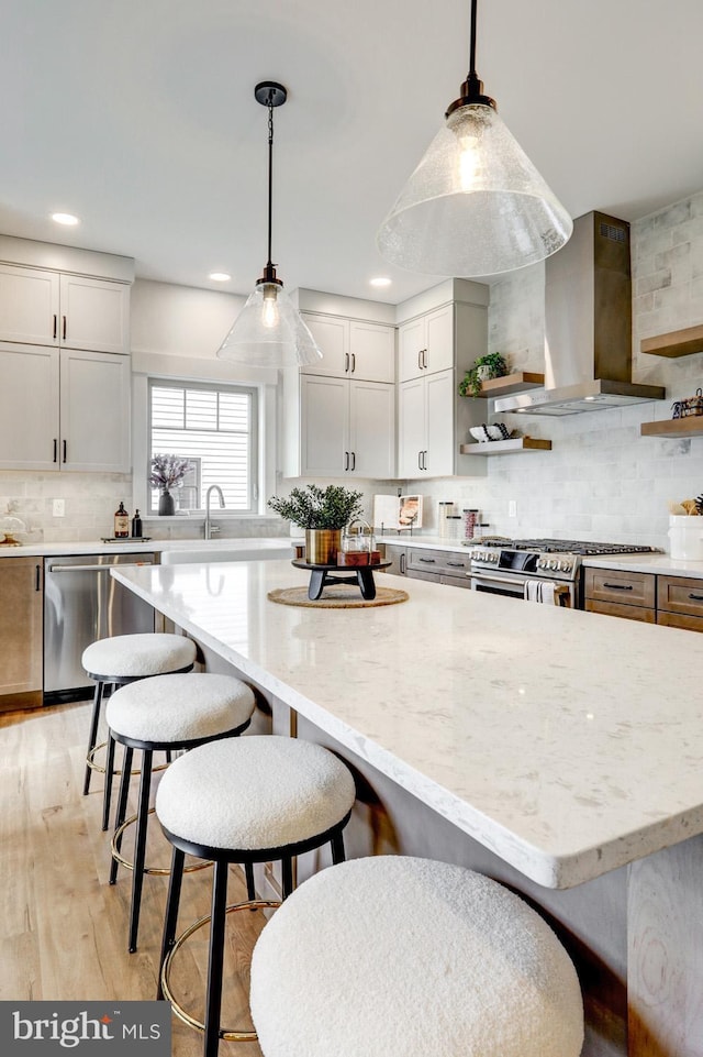 kitchen with appliances with stainless steel finishes, decorative light fixtures, white cabinetry, light stone counters, and wall chimney range hood