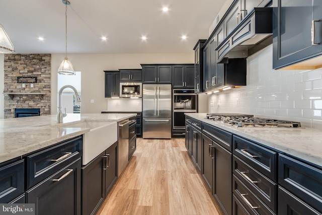 kitchen with a stone fireplace, light wood-type flooring, decorative light fixtures, light stone counters, and stainless steel appliances