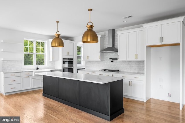 kitchen with wall chimney exhaust hood, light wood-type flooring, a kitchen island, white cabinetry, and stainless steel appliances