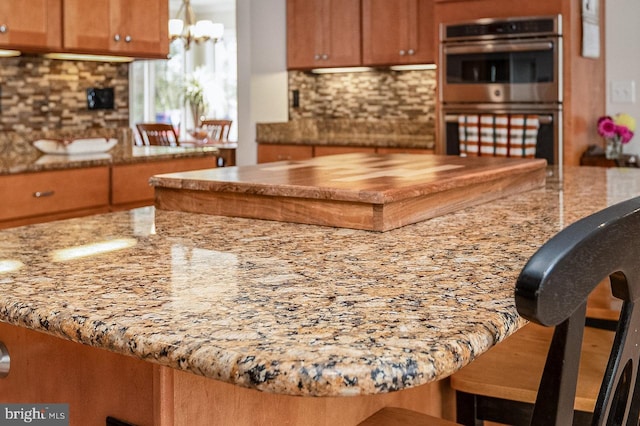 kitchen with a chandelier, stainless steel double oven, light stone countertops, and backsplash