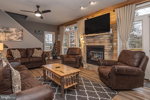 living room with ceiling fan, wood walls, a stone fireplace, and wood-type flooring