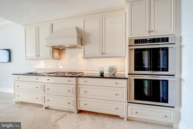 kitchen featuring premium range hood, cream cabinets, decorative backsplash, dark stone countertops, and stainless steel appliances