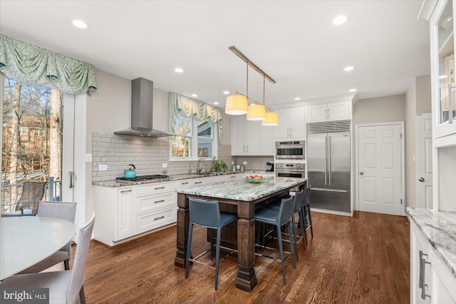 kitchen with a kitchen island, decorative light fixtures, light stone counters, stainless steel appliances, and wall chimney range hood