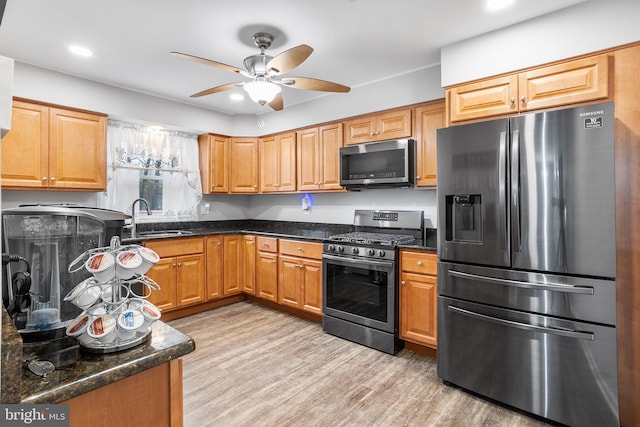 kitchen with appliances with stainless steel finishes, dark stone counters, ceiling fan, sink, and light hardwood / wood-style flooring