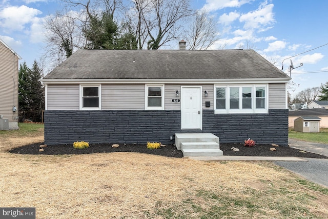 view of front of property featuring a storage shed, a shingled roof, stone siding, an outdoor structure, and central air condition unit