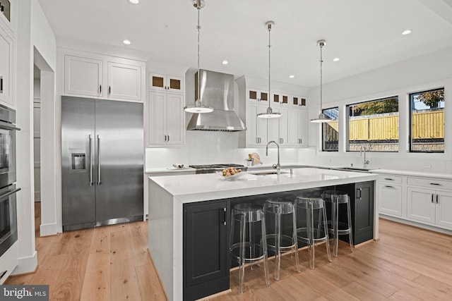 kitchen featuring wall chimney exhaust hood, an island with sink, light hardwood / wood-style floors, high end fridge, and white cabinets