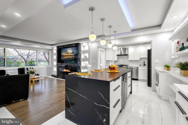 kitchen featuring a center island, wall chimney exhaust hood, stainless steel appliances, a tray ceiling, and white cabinets