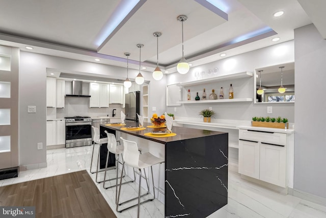 kitchen with white cabinetry, hanging light fixtures, stainless steel appliances, wall chimney range hood, and a raised ceiling