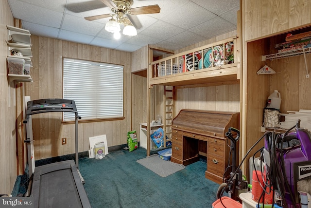 interior space with ceiling fan, a drop ceiling, dark carpet, and wood walls