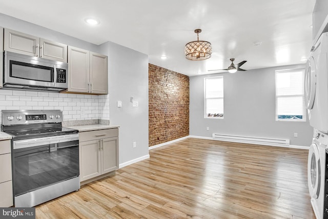 kitchen with gray cabinetry, a baseboard heating unit, light hardwood / wood-style floors, stacked washer and clothes dryer, and appliances with stainless steel finishes