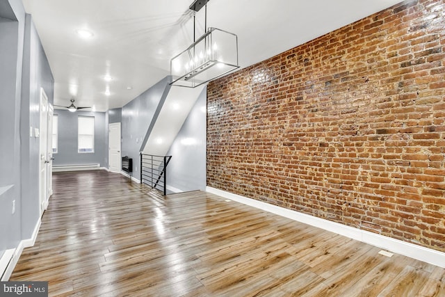 unfurnished living room featuring baseboard heating, ceiling fan, wood-type flooring, and brick wall