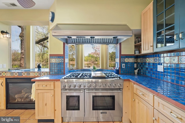 kitchen featuring backsplash, exhaust hood, range with two ovens, light brown cabinetry, and light tile patterned flooring