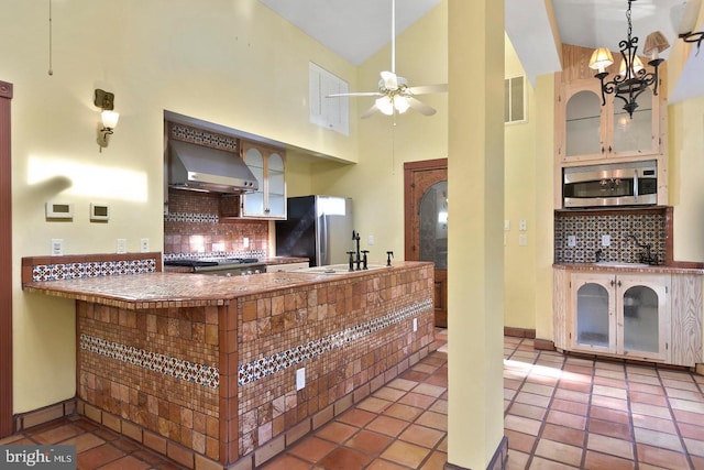 kitchen with backsplash, high vaulted ceiling, ceiling fan with notable chandelier, wall chimney range hood, and stainless steel appliances