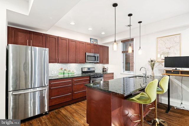 kitchen featuring backsplash, stainless steel appliances, dark wood-type flooring, a center island with sink, and hanging light fixtures