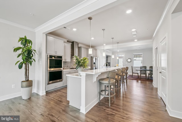 kitchen featuring wall chimney exhaust hood, hanging light fixtures, a breakfast bar, a center island with sink, and appliances with stainless steel finishes