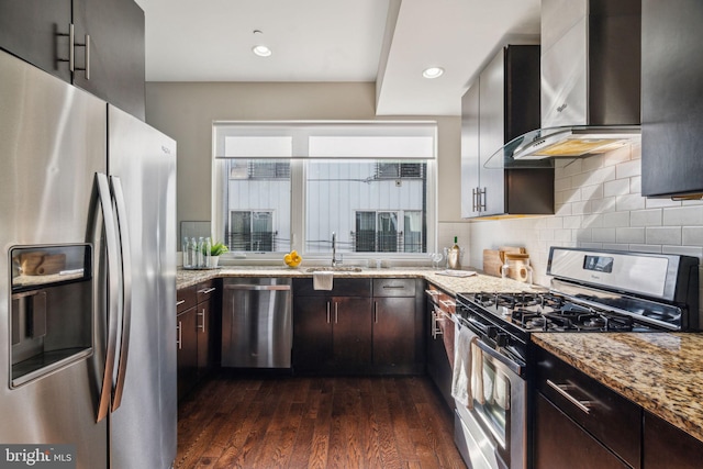 kitchen with appliances with stainless steel finishes, dark brown cabinets, wall chimney exhaust hood, dark wood-type flooring, and light stone countertops