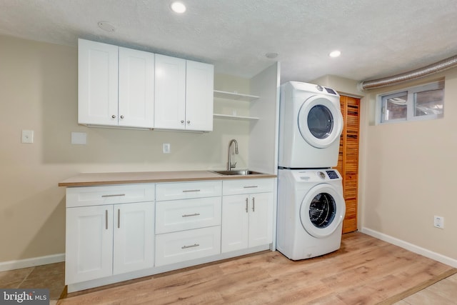 clothes washing area featuring stacked washing maching and dryer, sink, cabinets, a textured ceiling, and light hardwood / wood-style flooring