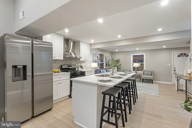 kitchen with stainless steel refrigerator with ice dispenser, wall chimney exhaust hood, gas range oven, white cabinetry, and an island with sink