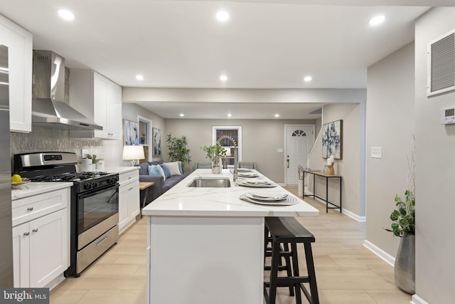 kitchen with white cabinets, stainless steel gas stove, wall chimney range hood, and a kitchen island with sink