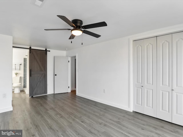 unfurnished bedroom featuring ensuite bath, hardwood / wood-style flooring, ceiling fan, a barn door, and a closet