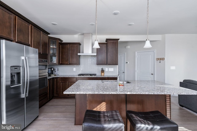 kitchen with hanging light fixtures, sink, light hardwood / wood-style flooring, wall chimney exhaust hood, and appliances with stainless steel finishes