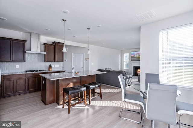 kitchen with light stone countertops, wall chimney exhaust hood, hanging light fixtures, light hardwood / wood-style floors, and a center island with sink